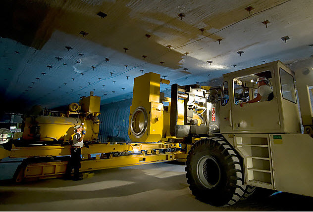 A heavy machinery vehicle sits in front of a piece of heavy equipment. A man in a hard hat drives the vehicle. A second man in a hard hat stands in front of the other machine and motions the driver toward him.