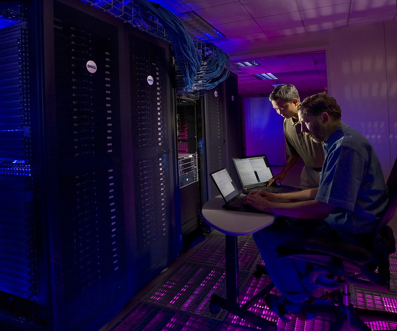 Two scientists look at laptop screens, facing Sandia's Thunderbird supercomputing cluster in a room filled with purple light.