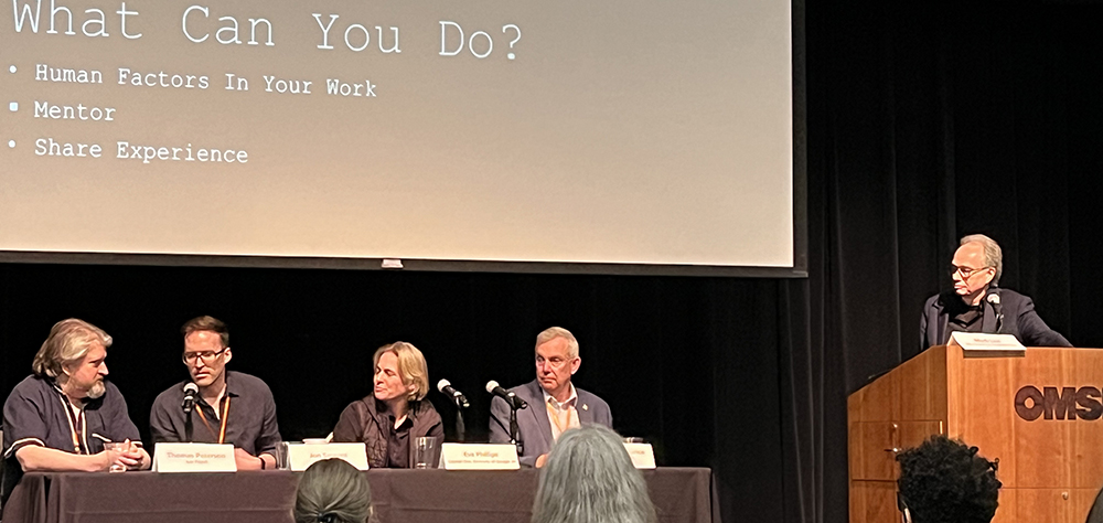 Four people sitting at a table in front of an audience with a projector screen above them. One person standing at a podium to the right.