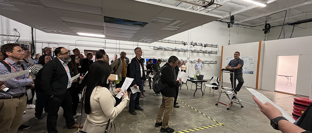 A large group of people standing in a room with a drop ceiling, with a man standing on a step ladder in front of them.
