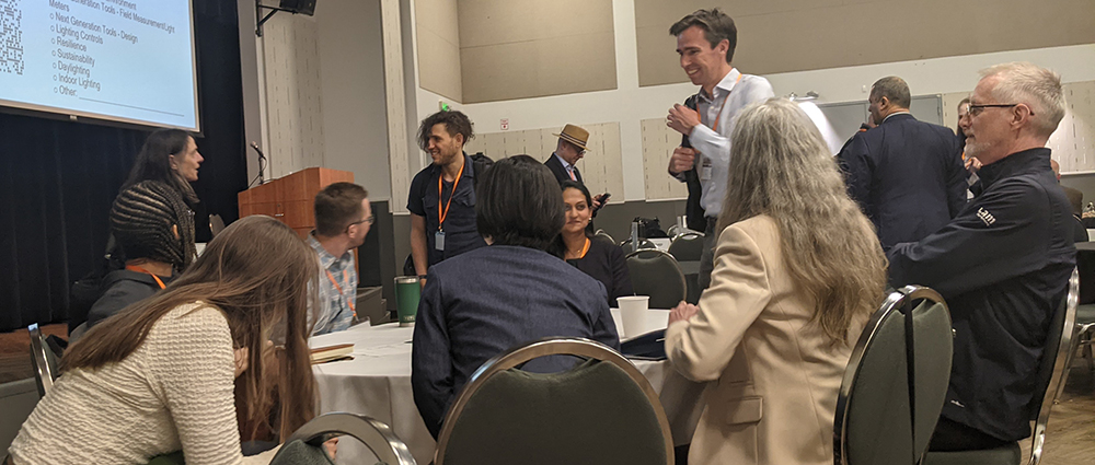 People sitting and standing around a round table having a discussion, with other people standing behind them.