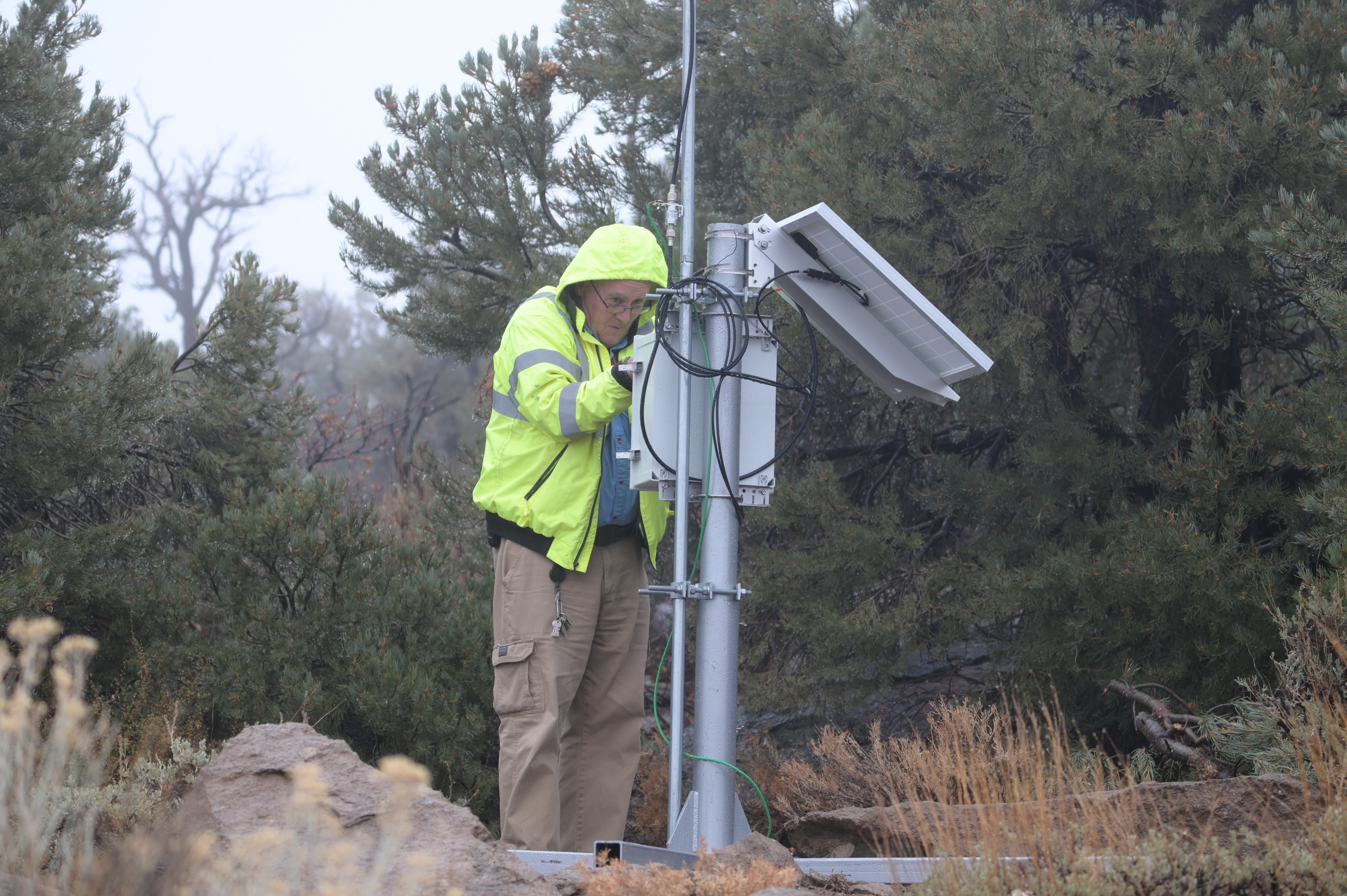 A man in a yellow jacket and tan pants stands and inspects equipment outdoors