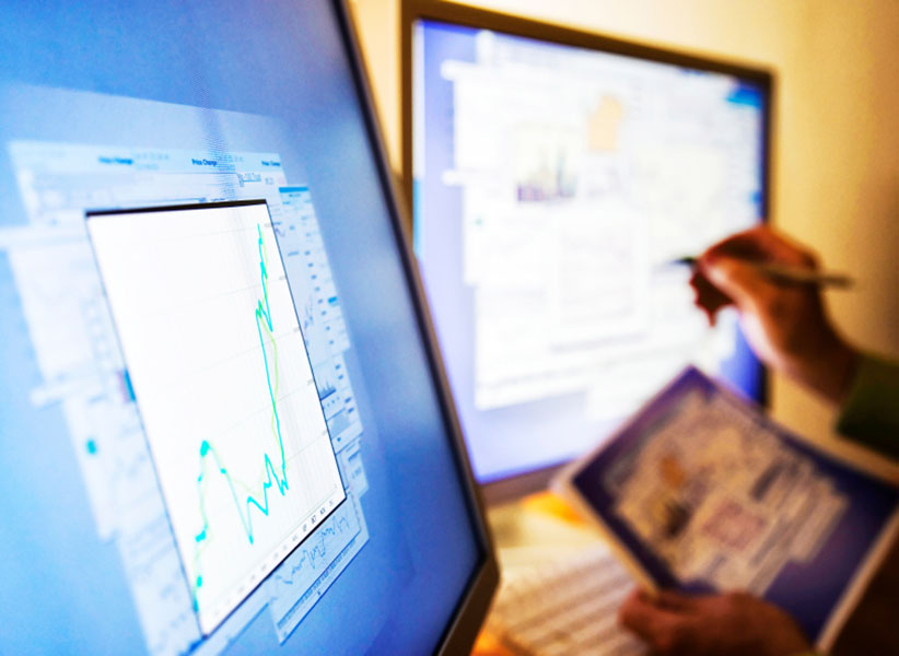 Two computer screens showing line and bar graphs. Someone is double-checking the graphs against information on a piece of paper.