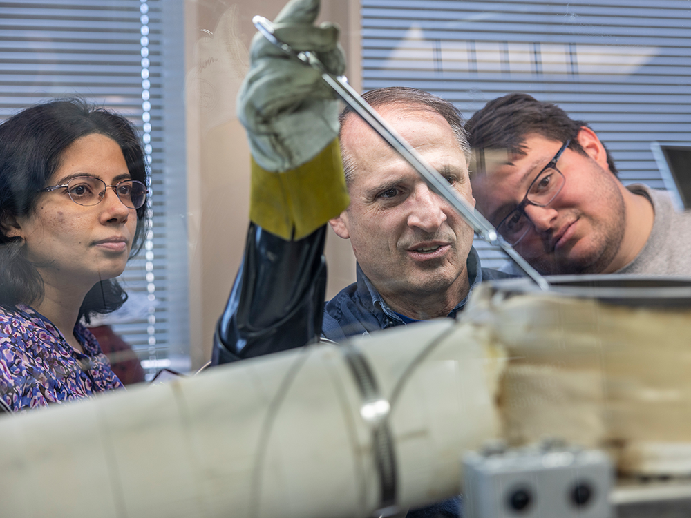 Three scientists watch as the center one holds a tool with a gloved hand.