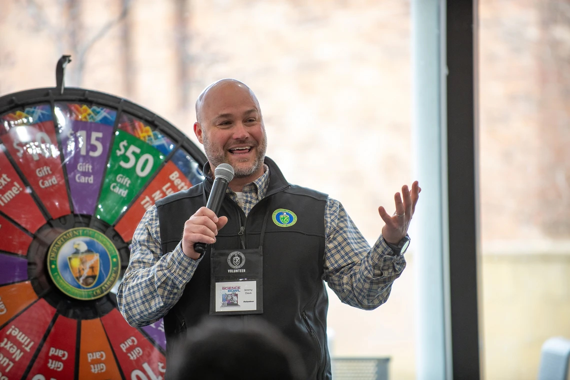 A man with a microphone talks to a crowd with a spin-to-win wheel behind him