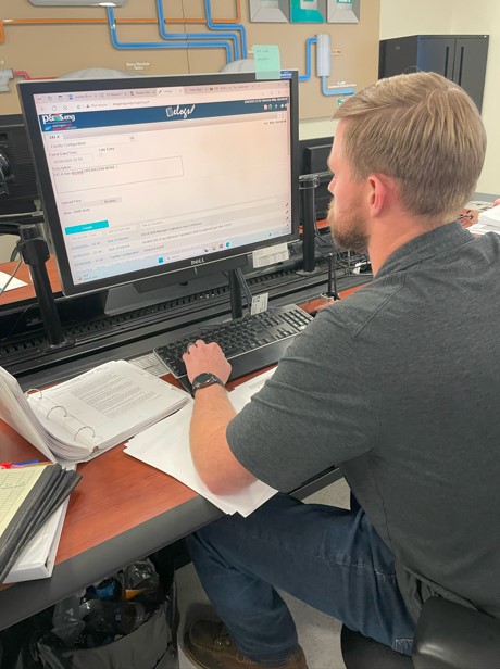 An employee sitting at a desk working on a computer with papers and binders on the desk