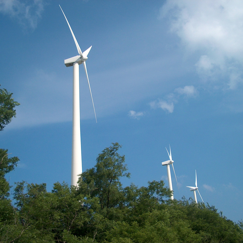 Three wind turbines behind a green tree line and against a blue sky with light, wispy clouds.