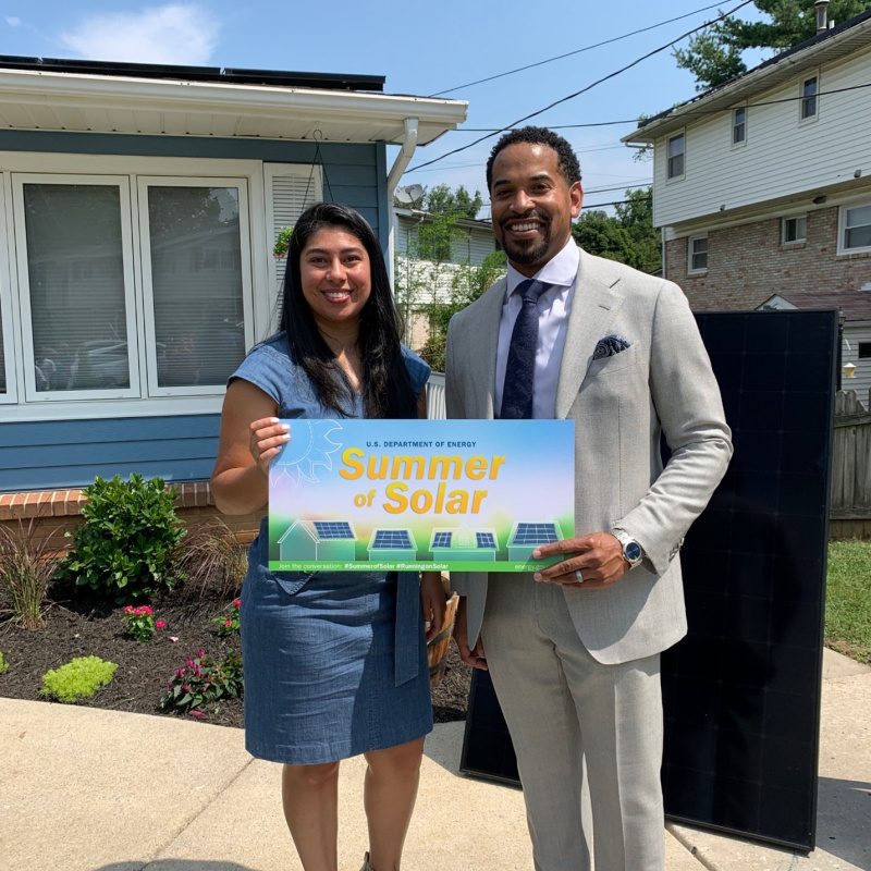 Two people standing in front of a home with solar panels holding a sign that says "Summer of Solar"
