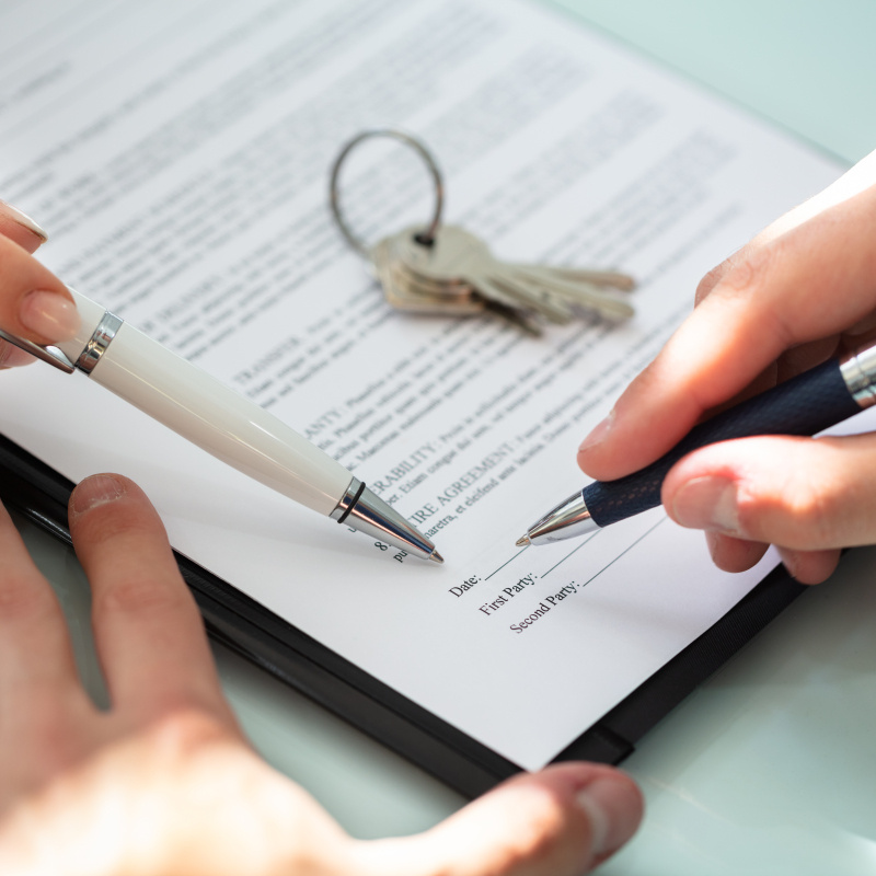 A contract on a table being signed by two people, each with a pen in hand, plus there is a set of keys sitting on top of the contract.