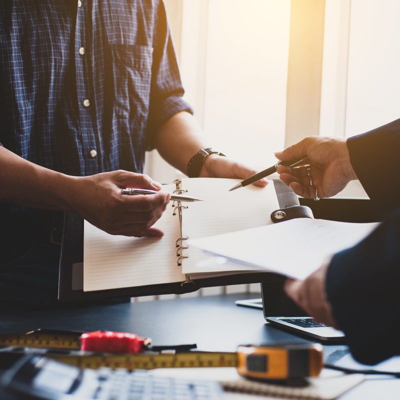 Partial view of the torsos of two men talking or having a meeting, each holding materials they're referring to.