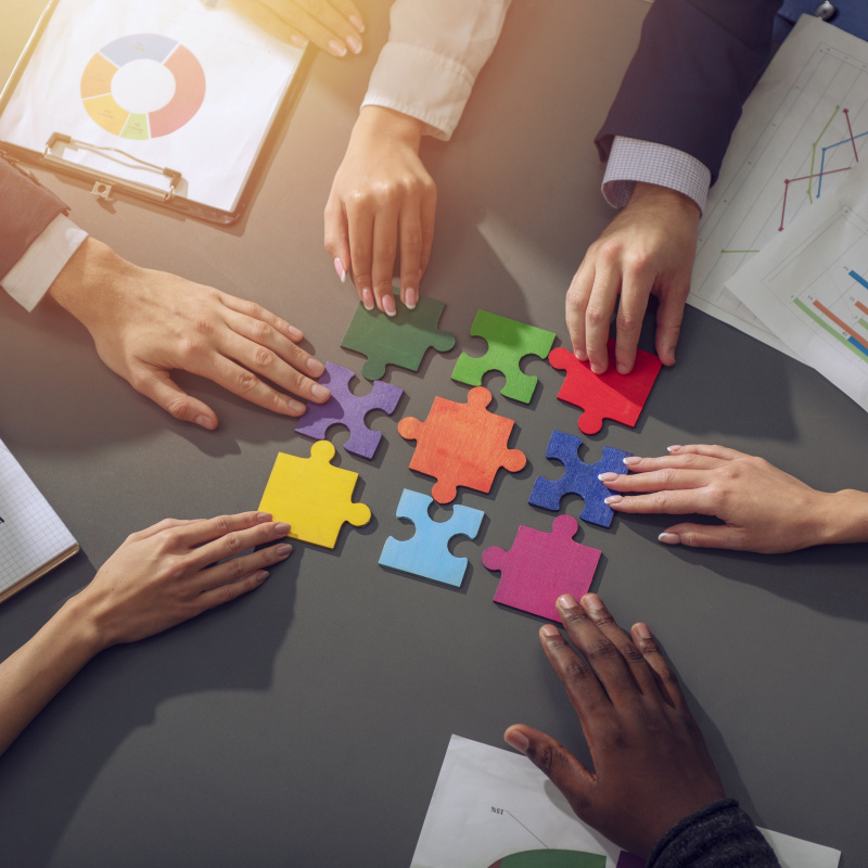 Large jigsaw puzzle pieces of different colors on a meeting table, with meeting attendee hands touching them.