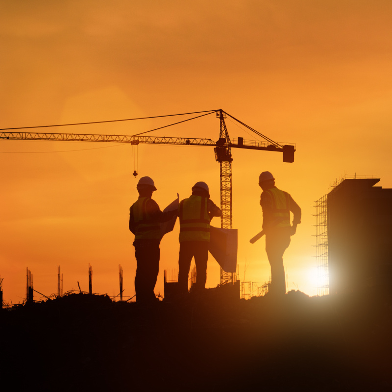 Silhouette image of engineers doing construction work with a tower crane and a setting sun in the background.