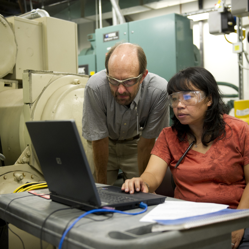 Two people wearing safety glasses looking at a laptop in a utility room.