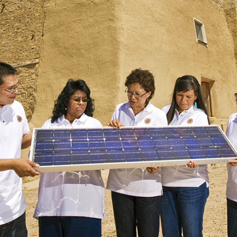 Five people wearing white shirts hold and look at a solar panel