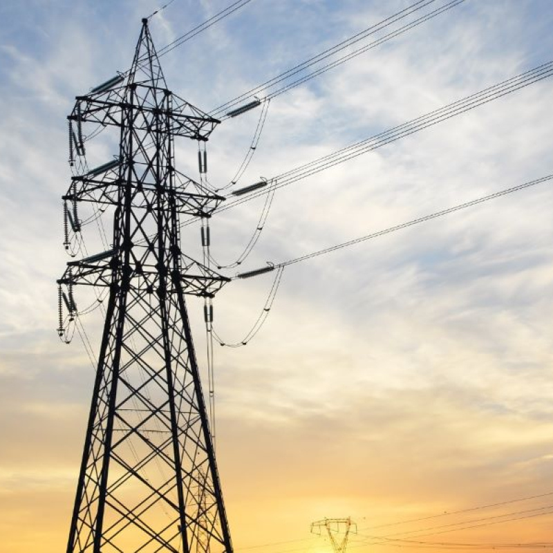 A large transmission tower and power lines appear in the foreground of the photo. In the background, the sun is setting in shades of yellow and gold against a cloudy light blue sky.