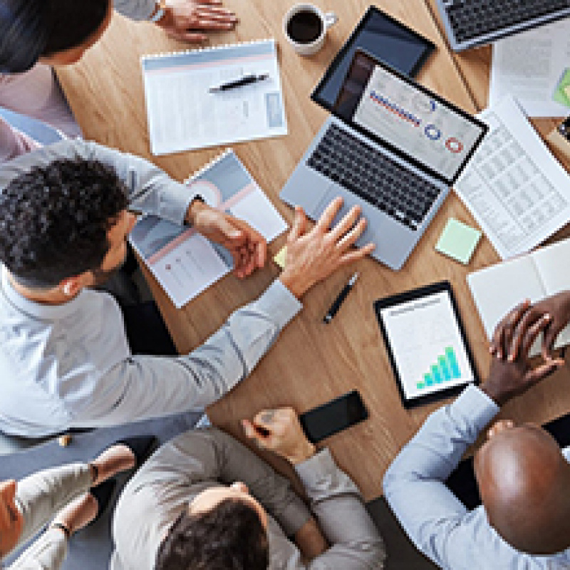 Photo looking down at a conference table surrounded by five people. On the table are laptops, tablets, and notebooks.