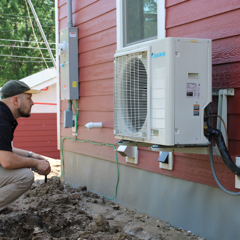 A crouching person inspects an air-source heat pump mounted to the side of a home.