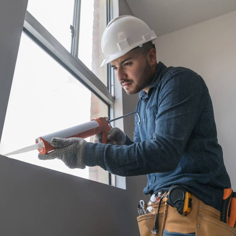 A person in a hard hat and applies caulk to a window.