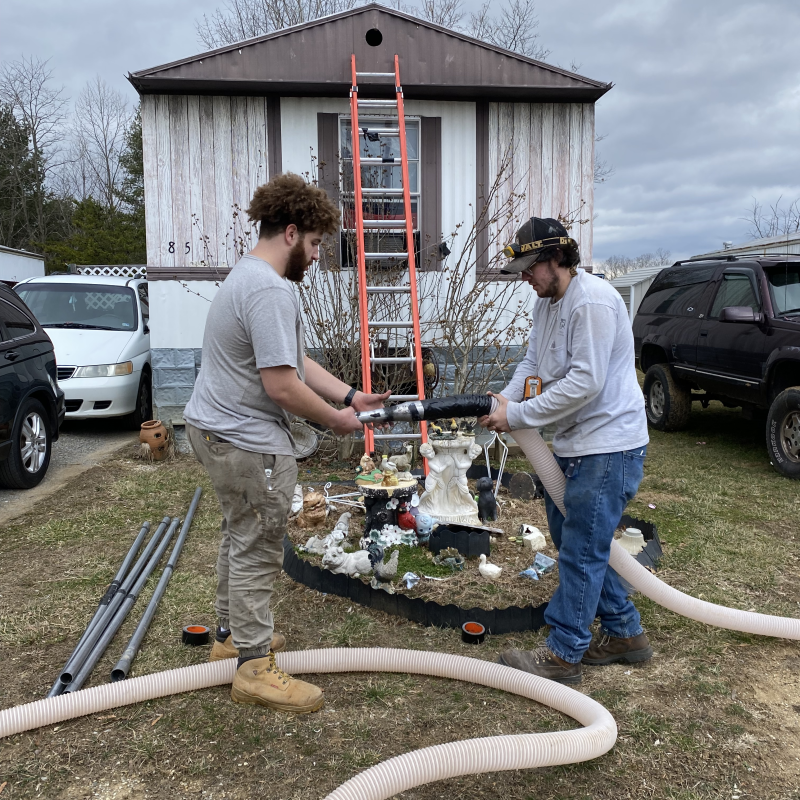 Two people hold a tube as they work in front of a mobile home.