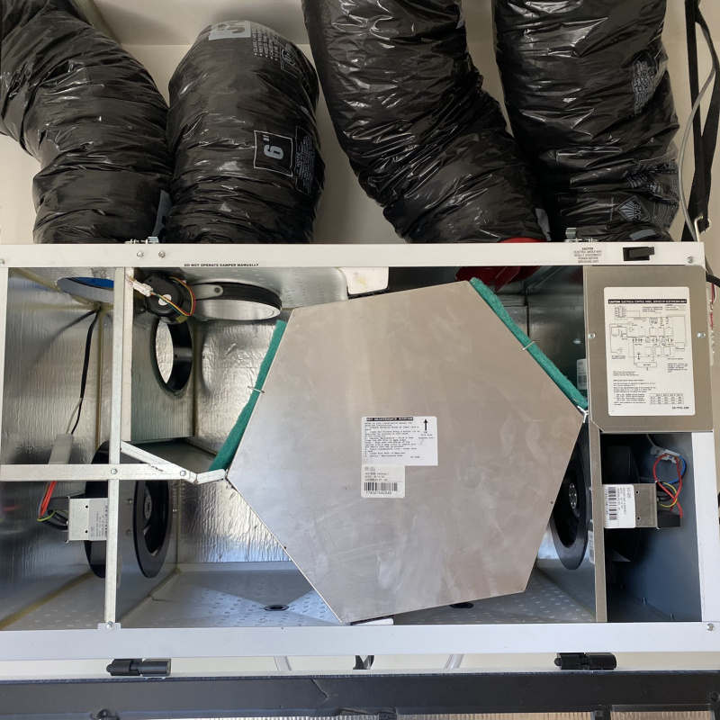 A piece of HVAC equipment mounted to a wall with exposed parts and four ducts connected to the top.