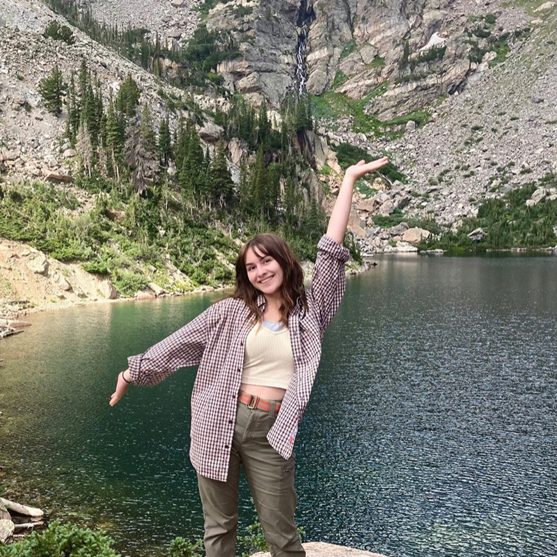 Person standing on rock with lake and rocky mountain in background.