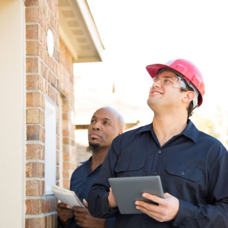 Two men, one wearing a hard hat, inspecting the exterior of a brick home.