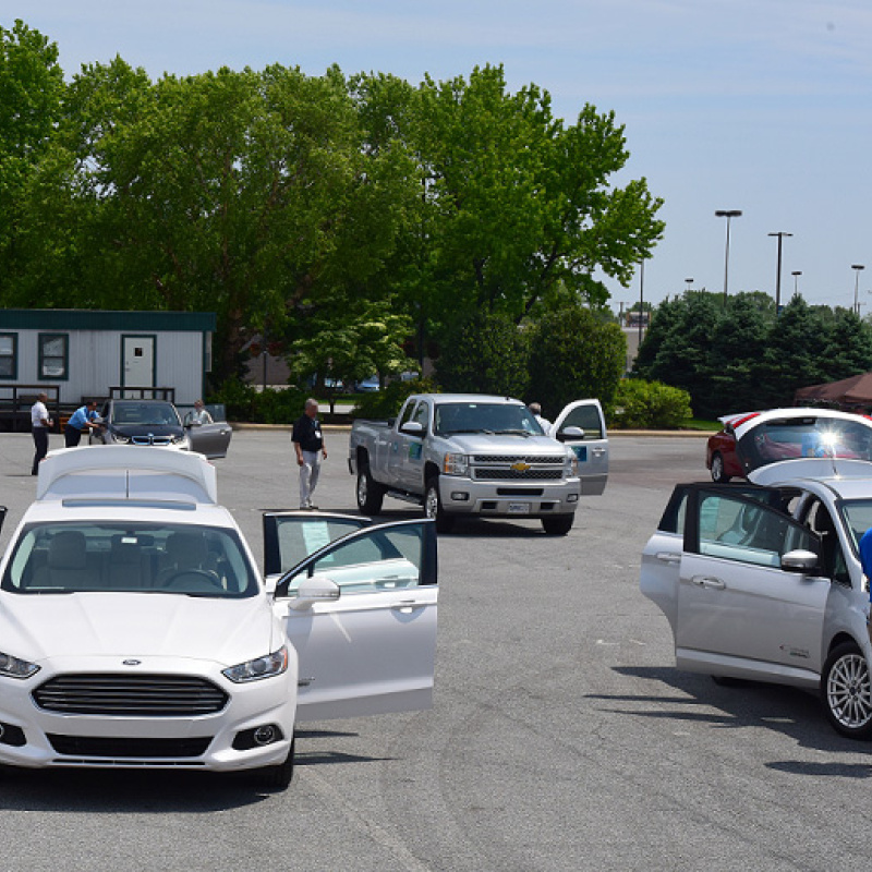 A variety of alternative fuel vehicles in a parking lot with several people looking at them.