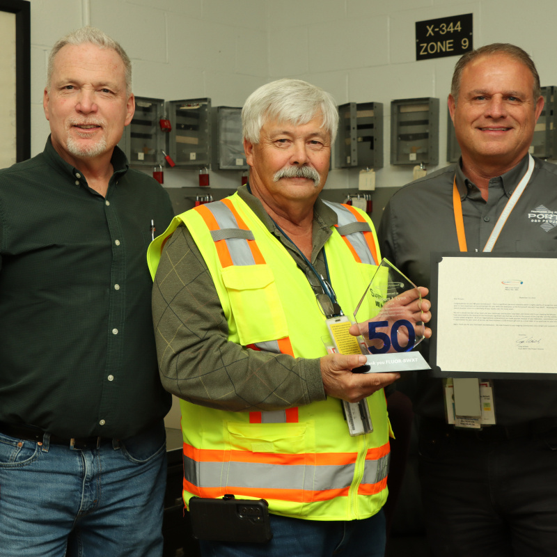 An employee at the Portsmouth Site in a yellow safety vest holding an award and posing for a group picture with two others