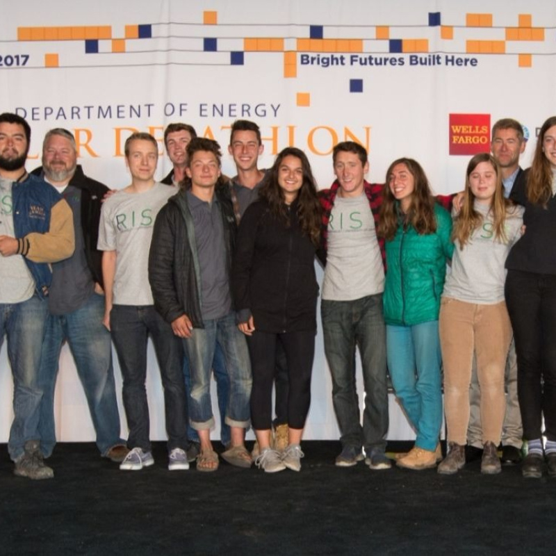 A group of Build Challenge student team members and organizers standing together, facing the camera, with a Solar Decathlon backdrop, and a lectern is nearby.