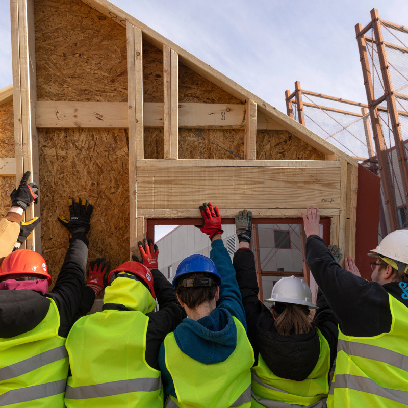 A crew of student workers in hard hats and construction vests, their backs to the camera, lifting a house structural panel in place.