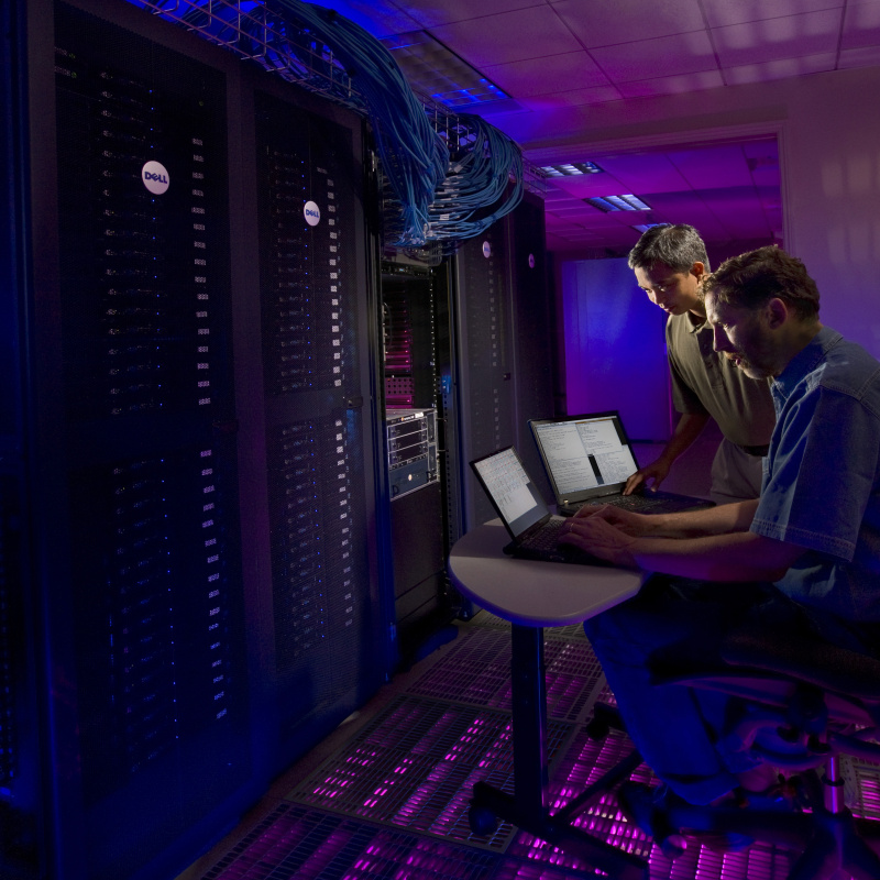 Two men operate a large collection of computers using laptops. 