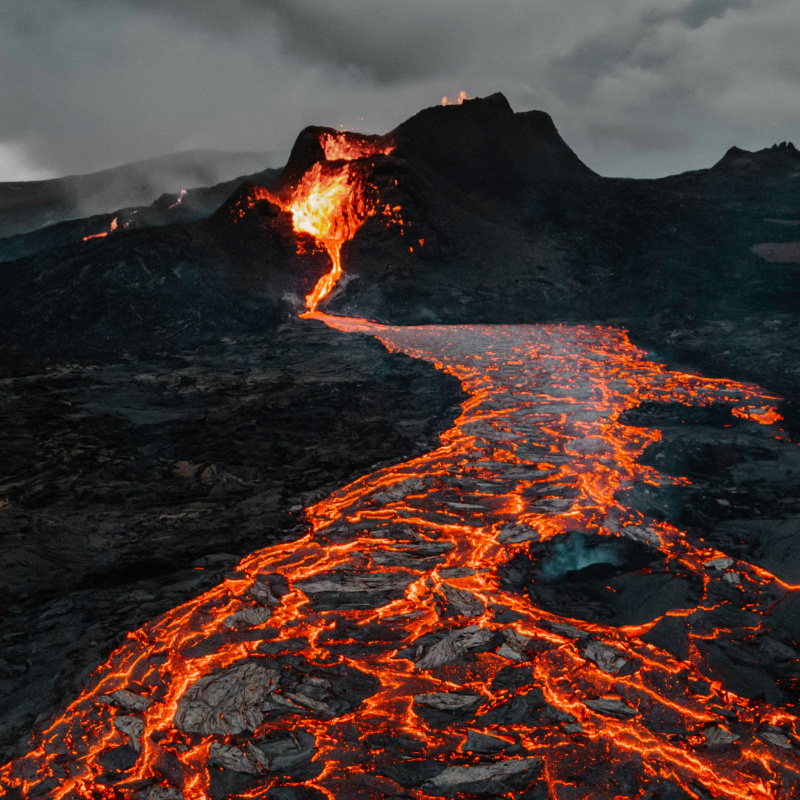 Photo of a small eruption with lava flowing away from it, against a dark rocky landscape with dark clouds in the sky.