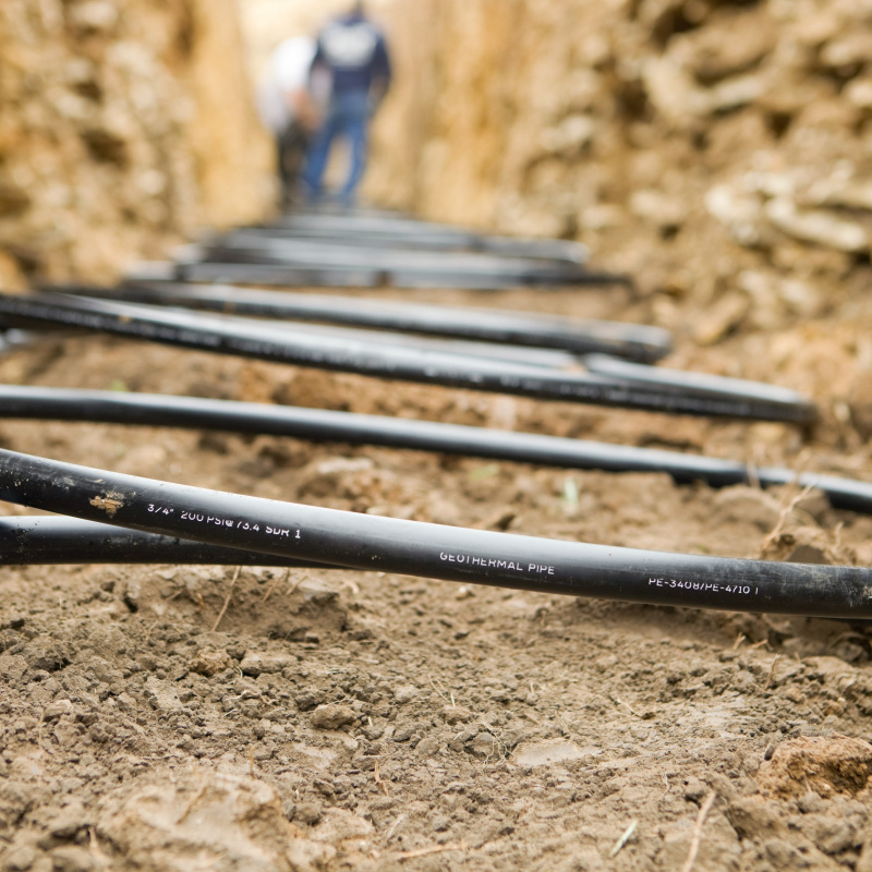 Close-up photo of thin, black plastic tubing in coils in approximately a five-foot-deep trench, with two people visible but out of focus at the far end of the ditch.