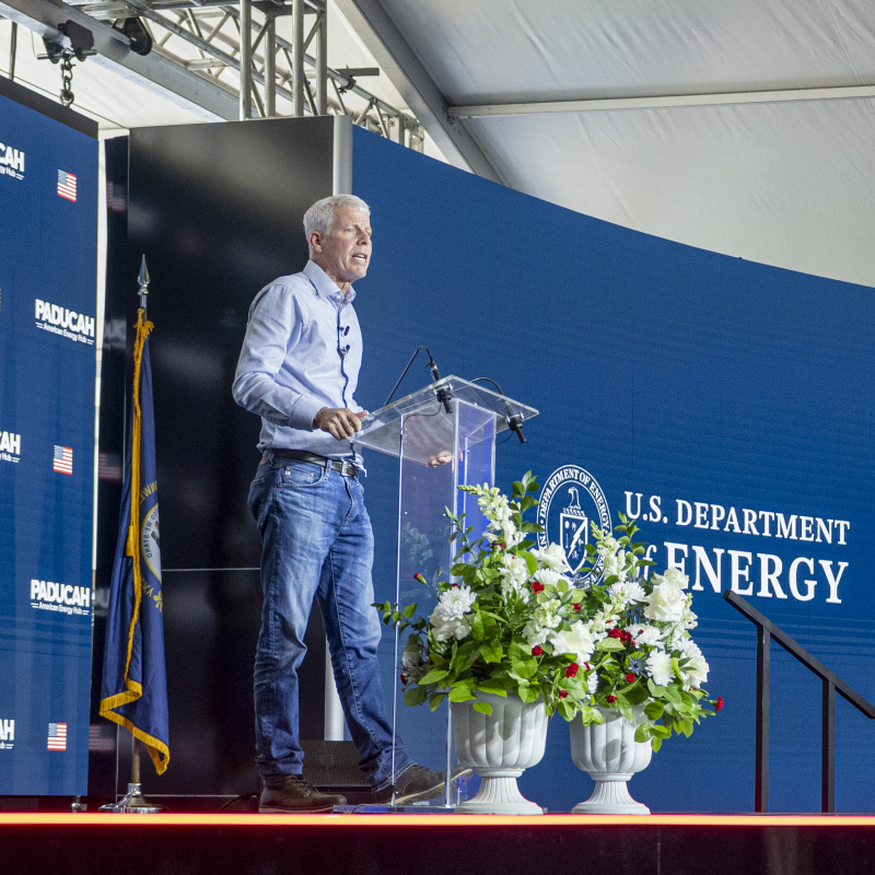 The US Department of Energy Secretary Chris Wright standing on a stage behind a clear podium with a blue backdrop behind him with the words Paducah Site and US Department of Energy written on the backdrops