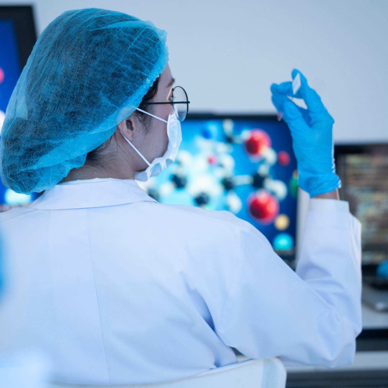 Laboratory researcher wearing protective gear examines a small sample, with molecular models displayed on computer screens.