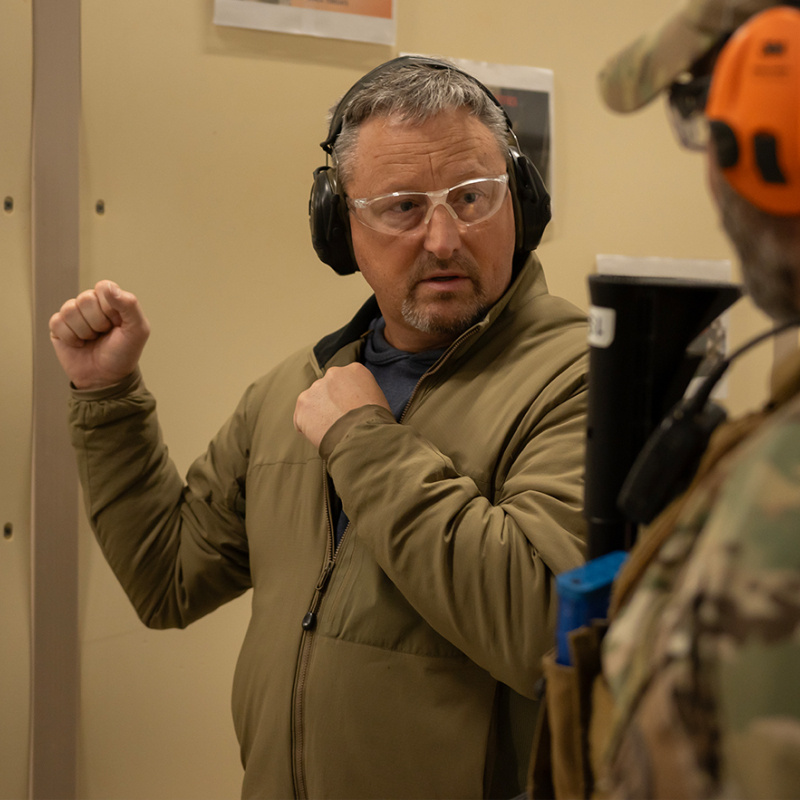 Two men at a firing range wearing camouflage speaking to each other 