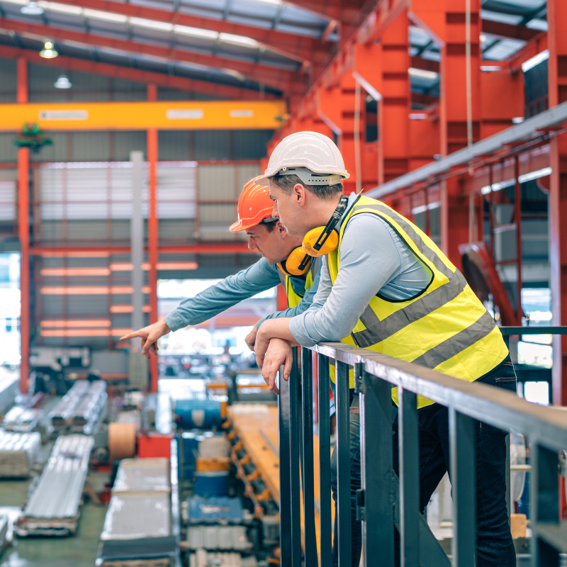 Two workers wearing hard hats and safety vests overlook operations inside a large manufacturing facility. 