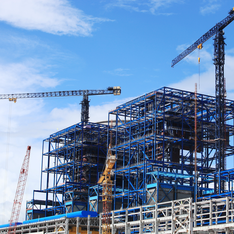 Large steel-frame building under construction with tower cranes against a blue sky. 
