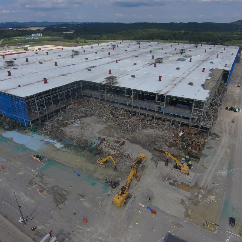 Aerial view of demolition taking place on the corner of a large building with a white roof and blue walls