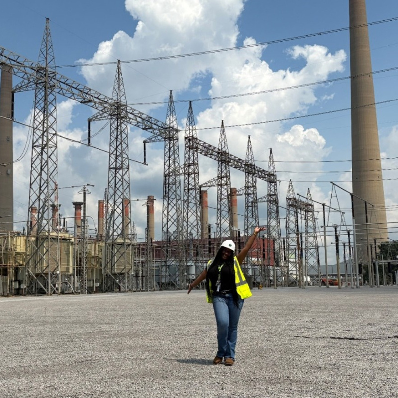 A woman in a white safety hat and yellow safety vest poses for a photo in front of tall electrical towers