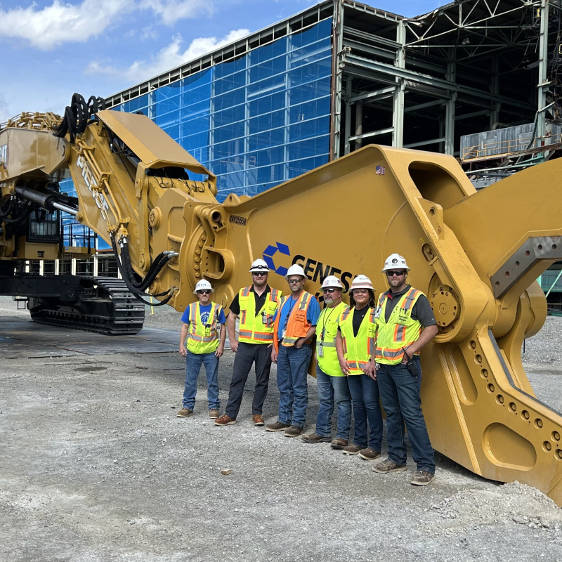 A group of people in safety gear standing beside a large yellow set of excavator shears