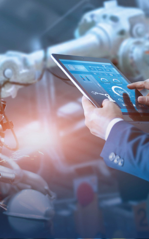 Photo of a person in a manufacturing factory, holding a digital device in front of industrial machinery.
