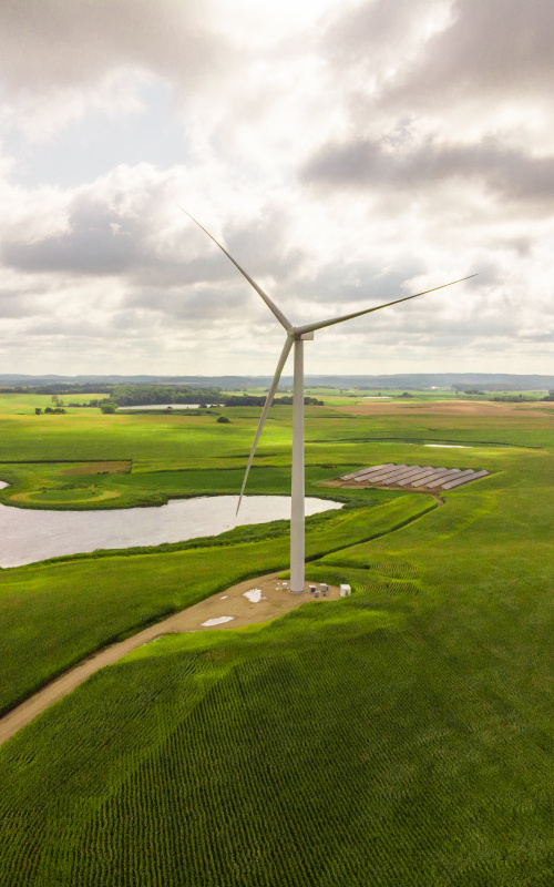 A large wind turbine on a grassy plain near a solar photovoltaic array and a lake.