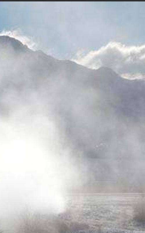 Photo of the steam eminating from a geyser in the foreground with a mountain range, clouds, and blue sky in the background.