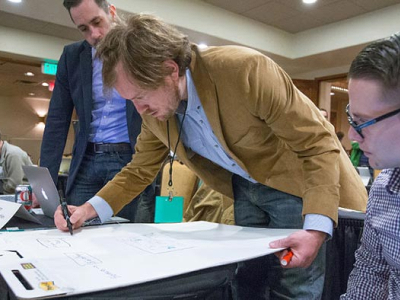 Image of three men working together at a table. The man in the middle writes on a large note pad while the other two men look on.