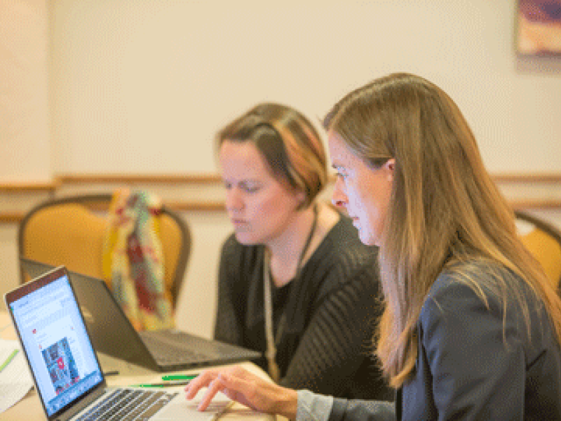 Image shows two women at a table each looking at their own laptop computers.
