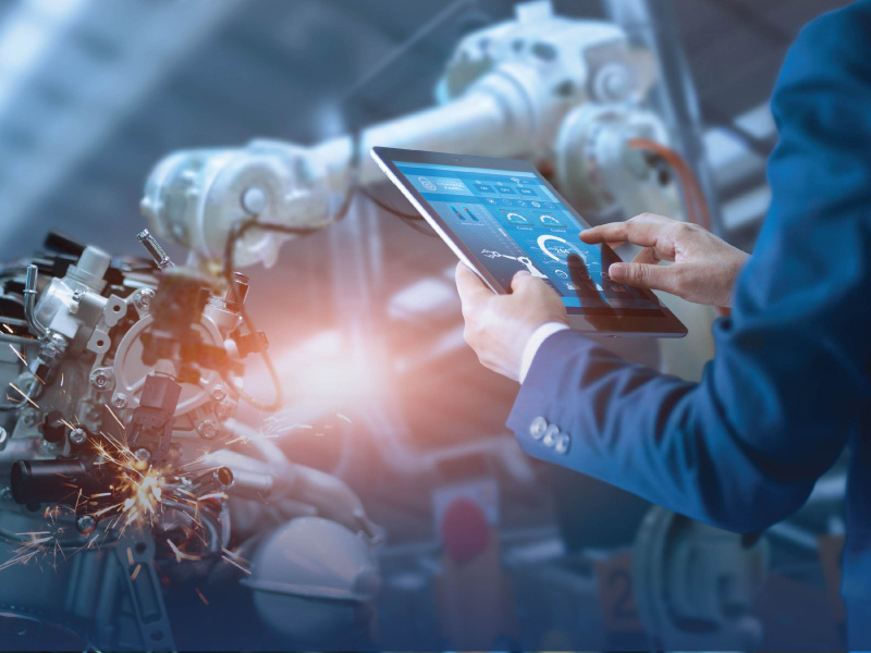 Photo of a person in a manufacturing factory, holding a digital device in front of industrial machinery.