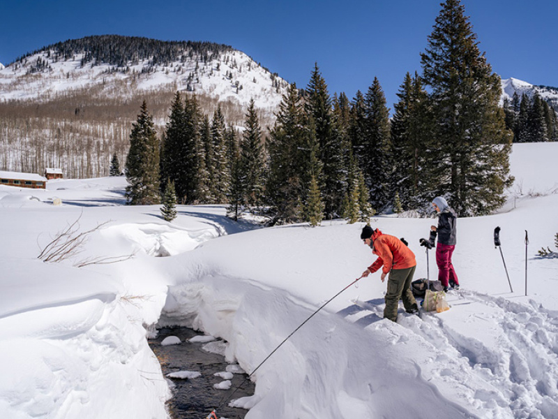 Two scientists in cold-weather clothing are standing in a snowy area next to a stream with mountains in the background. The one on the left is sticking a probe in the stream. There are hiking poles stuck in the snow behind both of them.