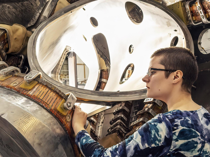 Alexandra LeViness (a white woman with a blue and white patterned long-sleeved shirt on), with her hand on a metal device much larger than her. A metal piece made of thick silver-colored metal with a ribbed copper-colored belt-like section snakes up and away from her, above her head.