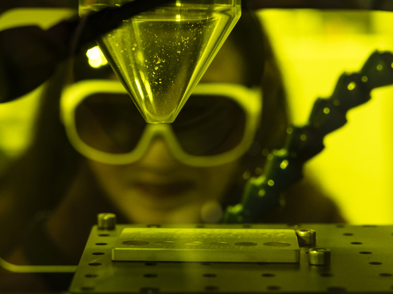 Madhavi Martin (a Southeast Asian woman with large lab goggles on) looks into a yellow-tinted box used for laser-induced breakdown spectroscopy. Inside the box is a clear funnel that is pointed towards a metal sheet with holes in it.
